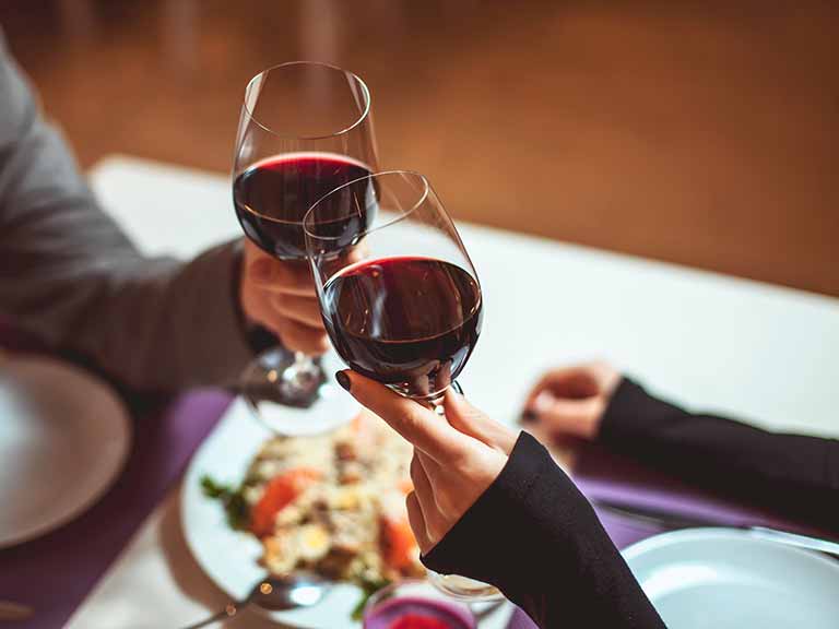 Two people toast with glasses of red wine over a meal at a table.