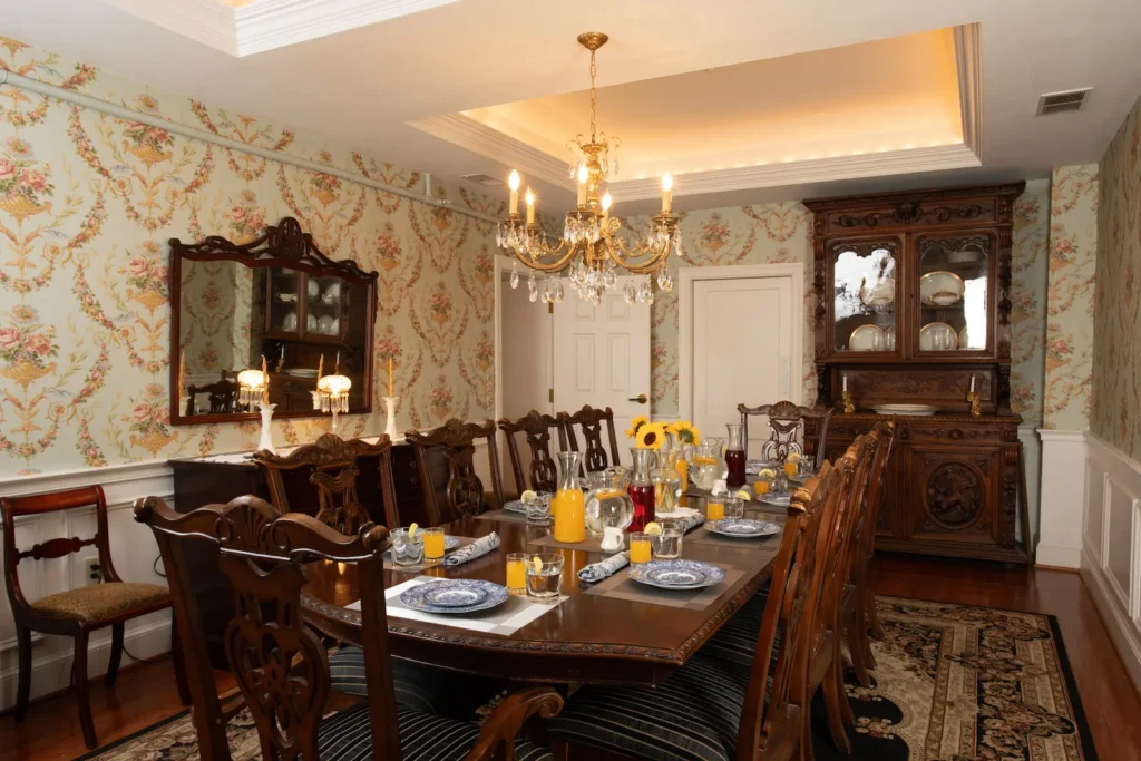 A formal dining room features a large wood table, chairs, wallpaper, and a crystal chandelier.
