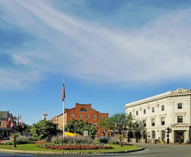 A city square with a roundabout, a central flag, a brick building, and a white stone building.